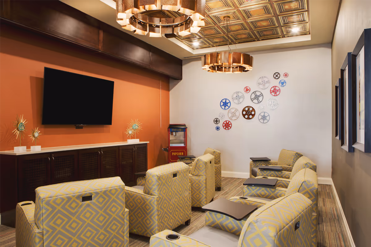 A cozy media room with multiple yellow patterned armchairs arranged in rows facing a large flat-screen TV mounted on an orange accent wall. Below the TV is a dark wooden cabinet with decorative starburst sculptures. A popcorn machine is placed in the corner, and the opposite wall features a decorative arrangement of colorful film reels. The ceiling has a decorative wooden panel design with two circular chandeliers.