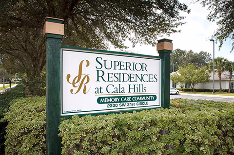 Outdoor sign for Superior Residences at Cala Hills, a memory care community located at 2300 SW 21st Circle, surrounded by greenery and trees with a parking lot and building in the background.
