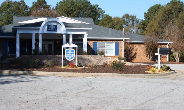 Exterior view of a single-story brick building with white columns and a dark roof, surrounded by trees and landscaping. There is a sign in front that reads 'Oxton Building B' and a mailbox on the right side near the driveway.