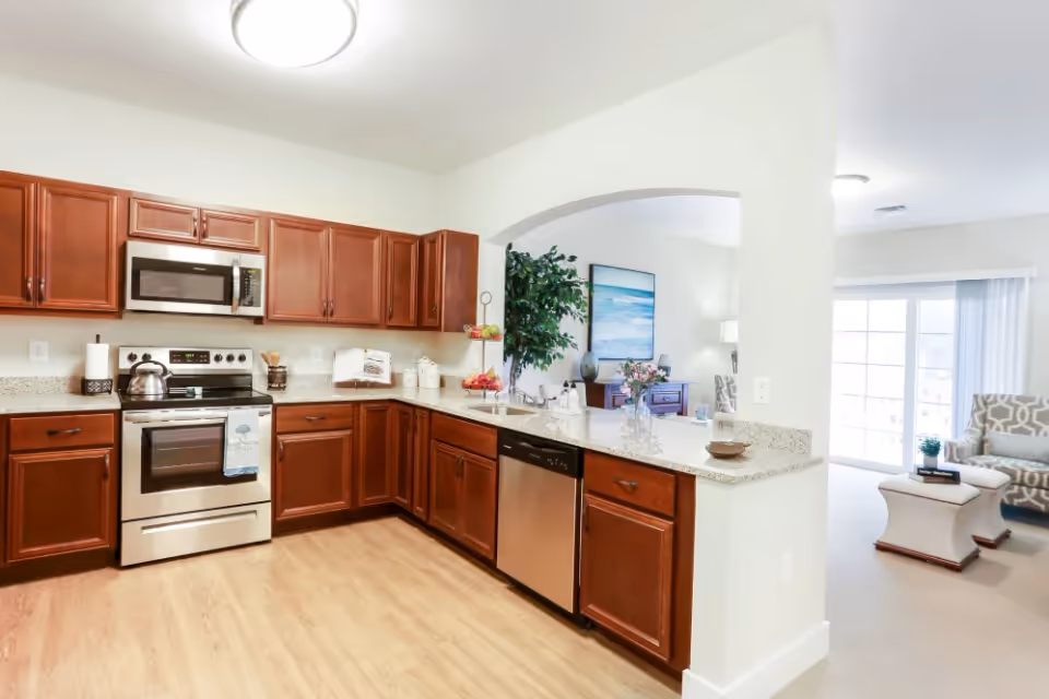 A modern kitchen with wooden cabinets, stainless steel appliances including a microwave, oven, and dishwasher. The kitchen has granite countertops and a light wood floor. There is an open archway leading to a living room area with a large window or sliding glass door letting in natural light. The living room has a patterned armchair, a small table, and a large plant near the archway.