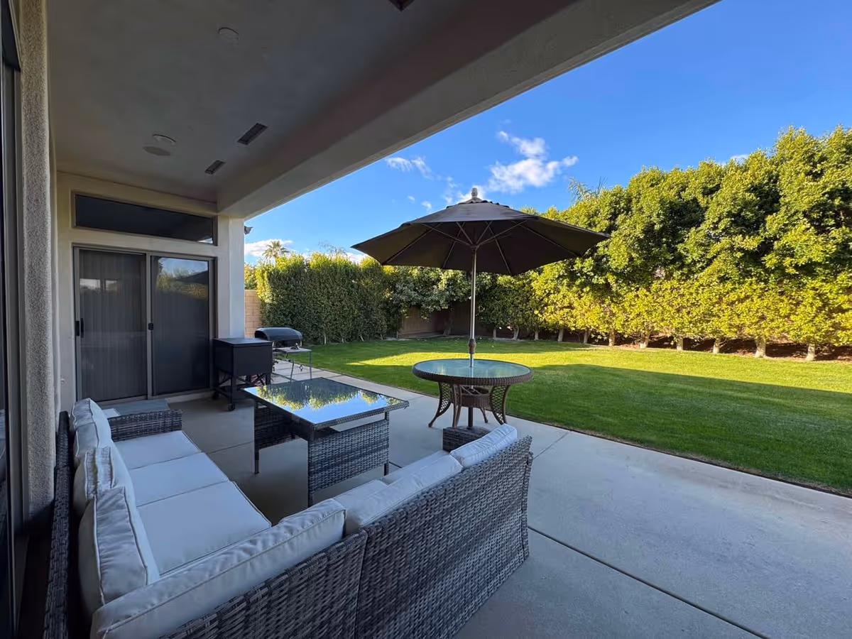Covered patio area with a wicker sectional sofa with white cushions, a glass-top coffee table, and a round table with a large umbrella. The patio overlooks a well-maintained green lawn bordered by tall, dense bushes under a clear blue sky.