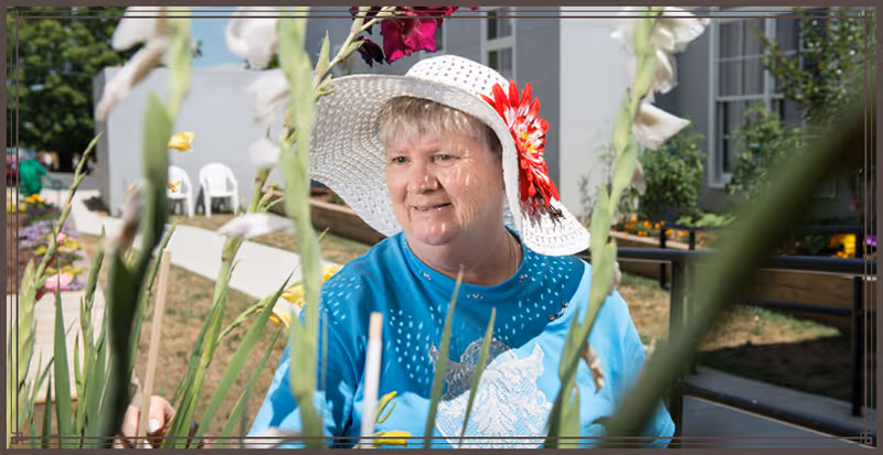 An elderly woman wearing a white sun hat with a red flower and a blue shirt is tending to tall flowers in an outdoor garden area with a building and white chairs in the background.