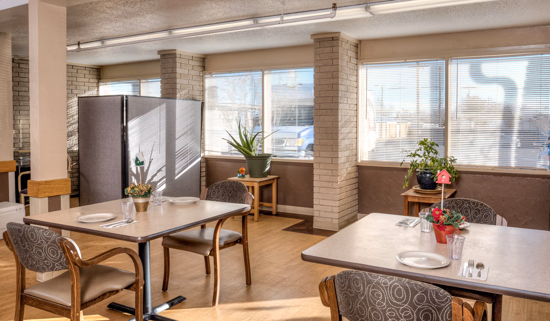 A bright dining area with two tables set with plates, glasses, and utensils. Each table has chairs with patterned upholstery. There are large windows with blinds letting in natural light, and small plants are placed on side tables near the windows. The room has light-colored brick pillars and wooden flooring.