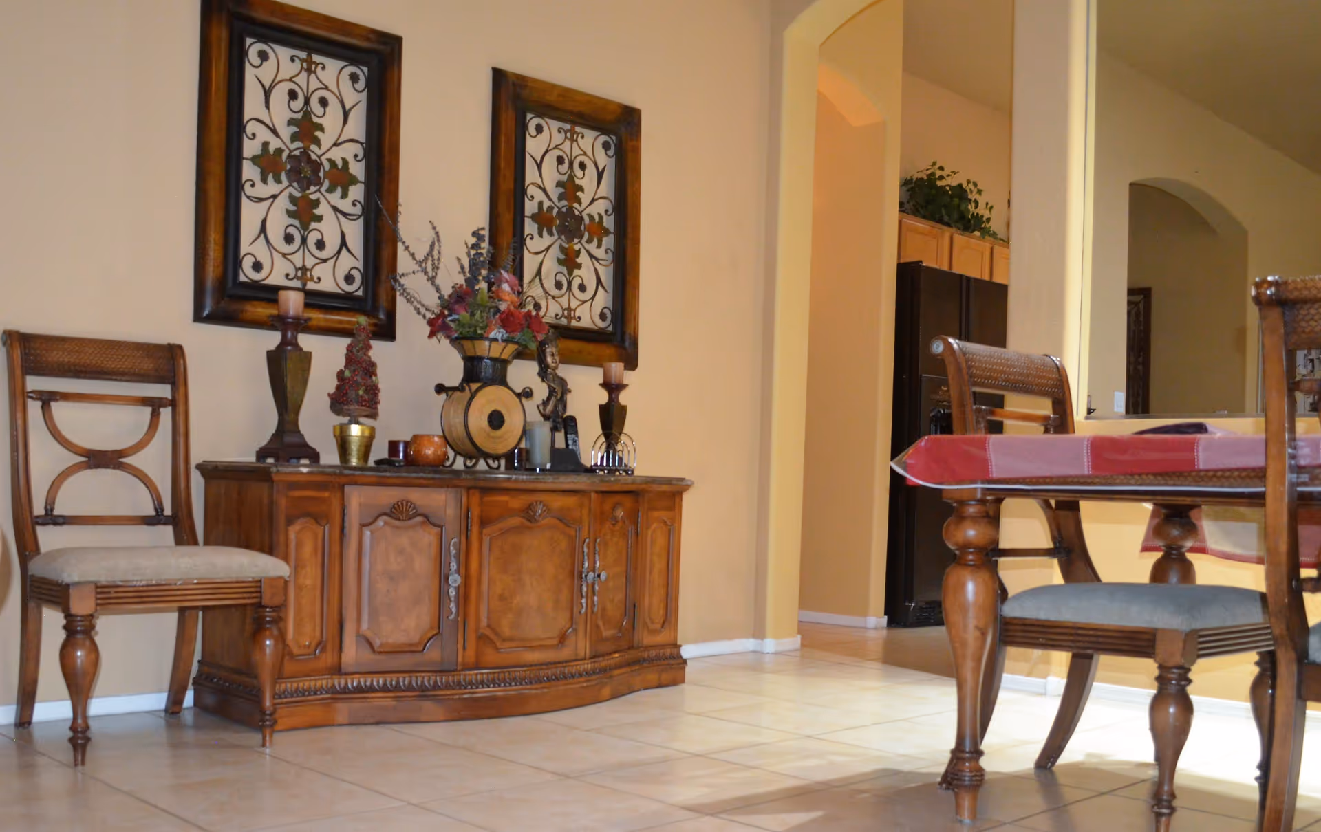 Interior view of a dining area with a wooden sideboard decorated with candles, a vase with flowers, and ornamental items. Two framed decorative wall hangings are above the sideboard. Part of a dining table with a red tablecloth and wooden chairs is visible on the right. The background shows an open doorway leading to a kitchen area with wooden cabinets and a black refrigerator.