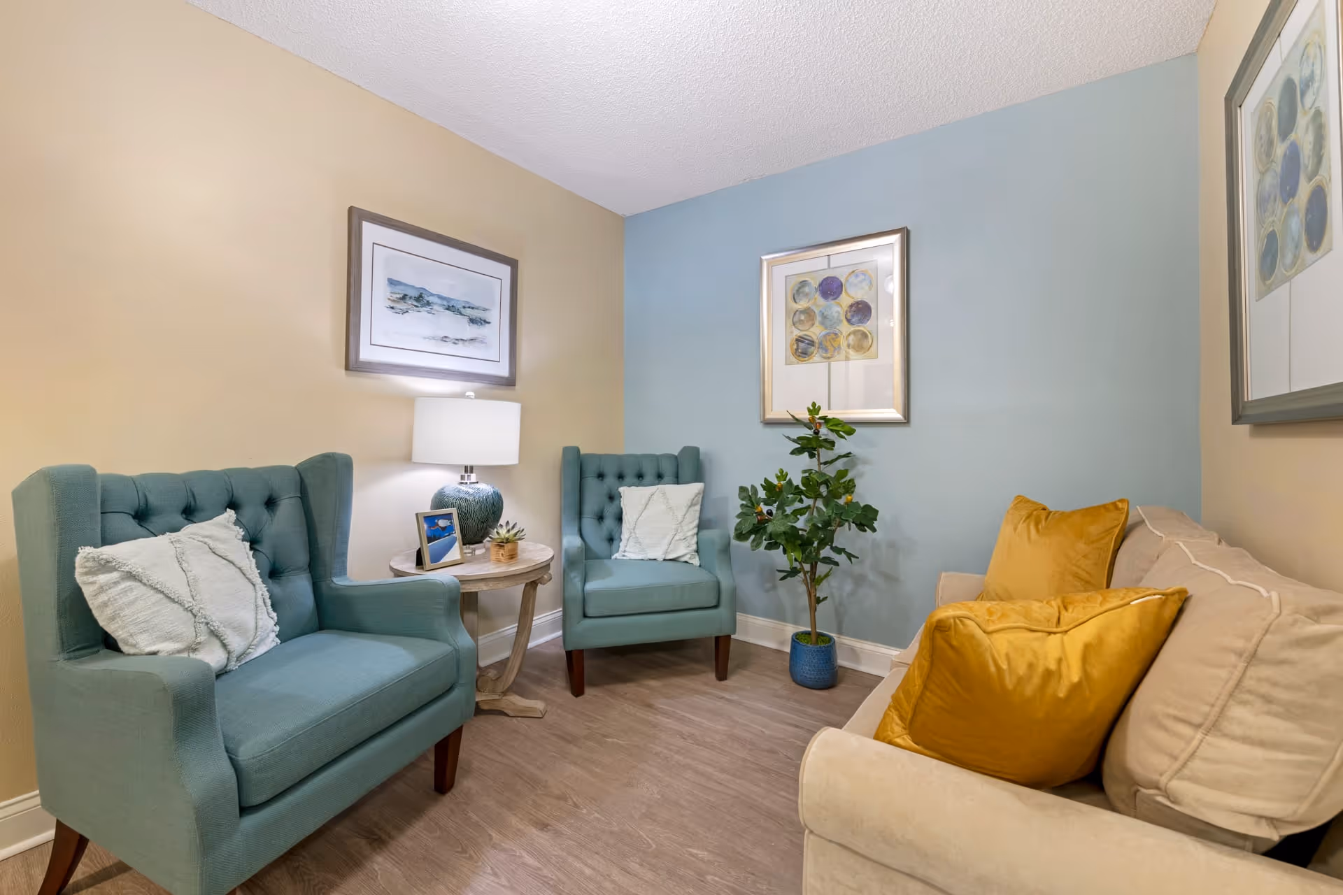 A cozy living room area with two teal armchairs, a beige sofa with mustard yellow pillows, a small round wooden side table with a lamp, a framed photo, and a small plant. The walls are painted in beige and light blue, adorned with framed artwork, and there is a potted plant in the corner.