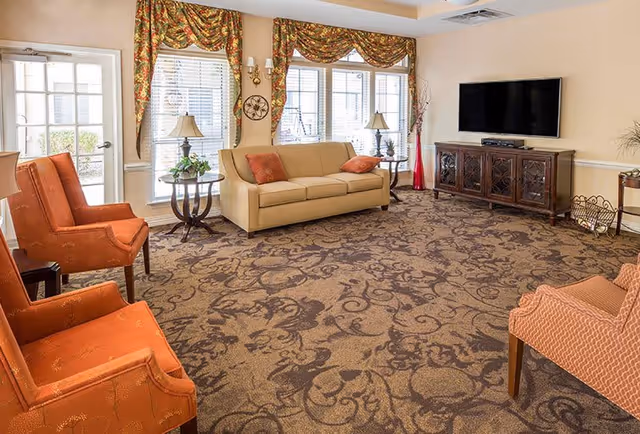 A cozy living room with a beige sofa and orange armchairs arranged around a patterned carpet. The room has large windows with floral valances, a flat-screen TV mounted on the wall above a wooden cabinet, and decorative lamps and plants on side tables.