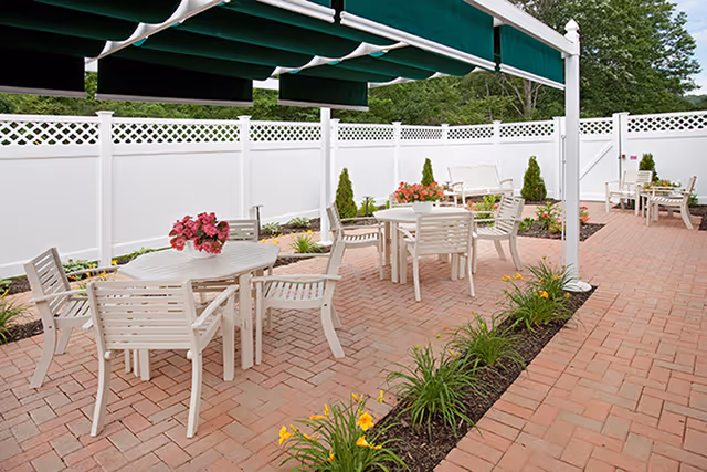 Outdoor brick patio with white tables and chairs under green awnings beside a white privacy fence and planted flower beds.