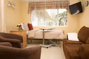 A cozy senior living room with a hospital-style bed near a large window covered by vertical blinds and a plaid valance. The room includes a brown sofa, two brown armchairs, a wooden side table with a flower vase, and a wall-mounted flat-screen TV.