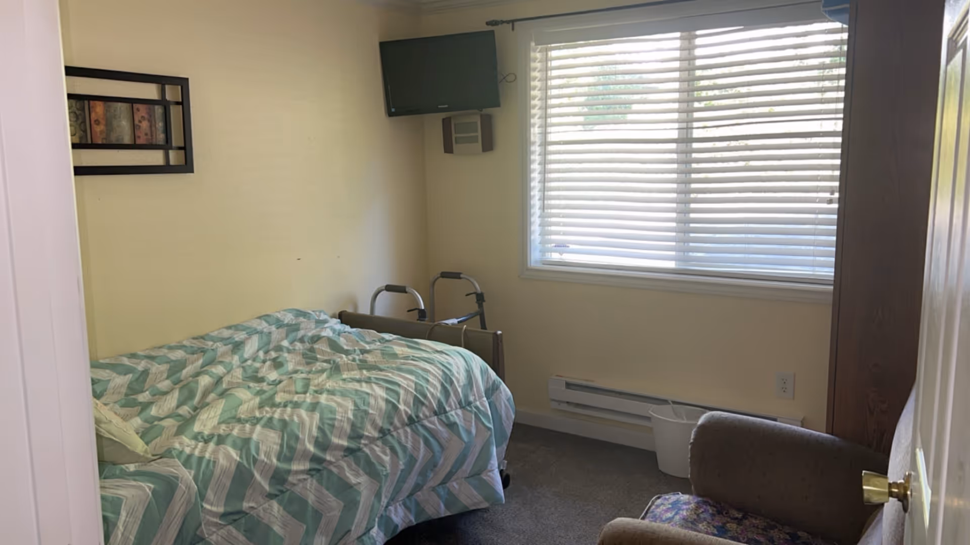 A small bedroom with a bed covered in a green and white chevron patterned comforter, a walker positioned at the foot of the bed, a wall-mounted TV above the bed, a window with white blinds letting in natural light, a beige armchair with a floral cushion, and a wastebasket near the window.