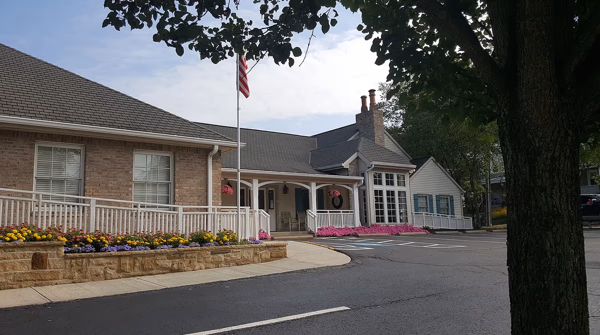 Exterior view of a single-story brick and siding building with a covered entrance, white railings, flower beds with colorful flowers, an American flag on a flagpole, and a large tree in the foreground.