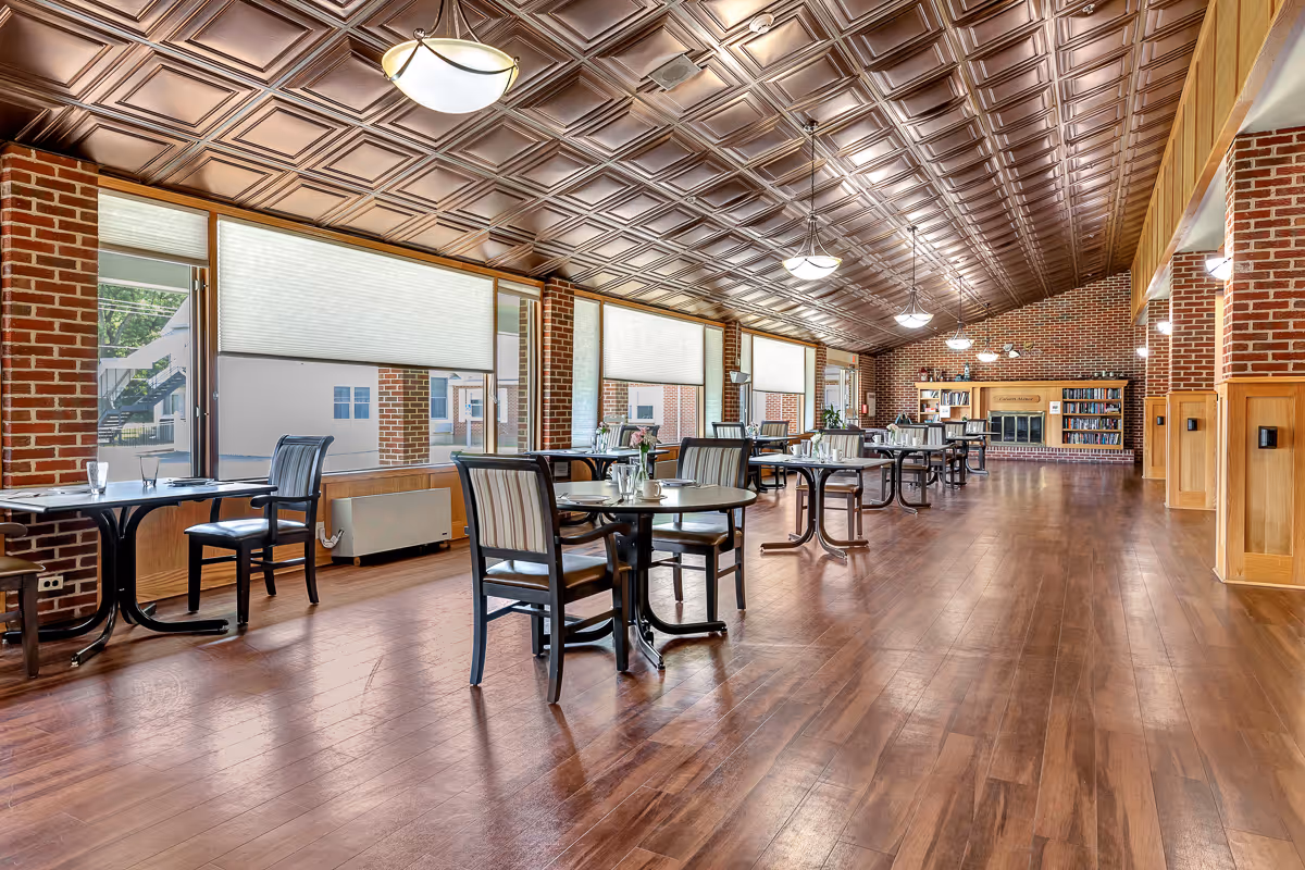 A spacious dining room with wooden floors, multiple tables and chairs arranged neatly, large windows with blinds, brick walls, and a decorative ceiling. At the far end, there is a fireplace flanked by bookshelves filled with books.