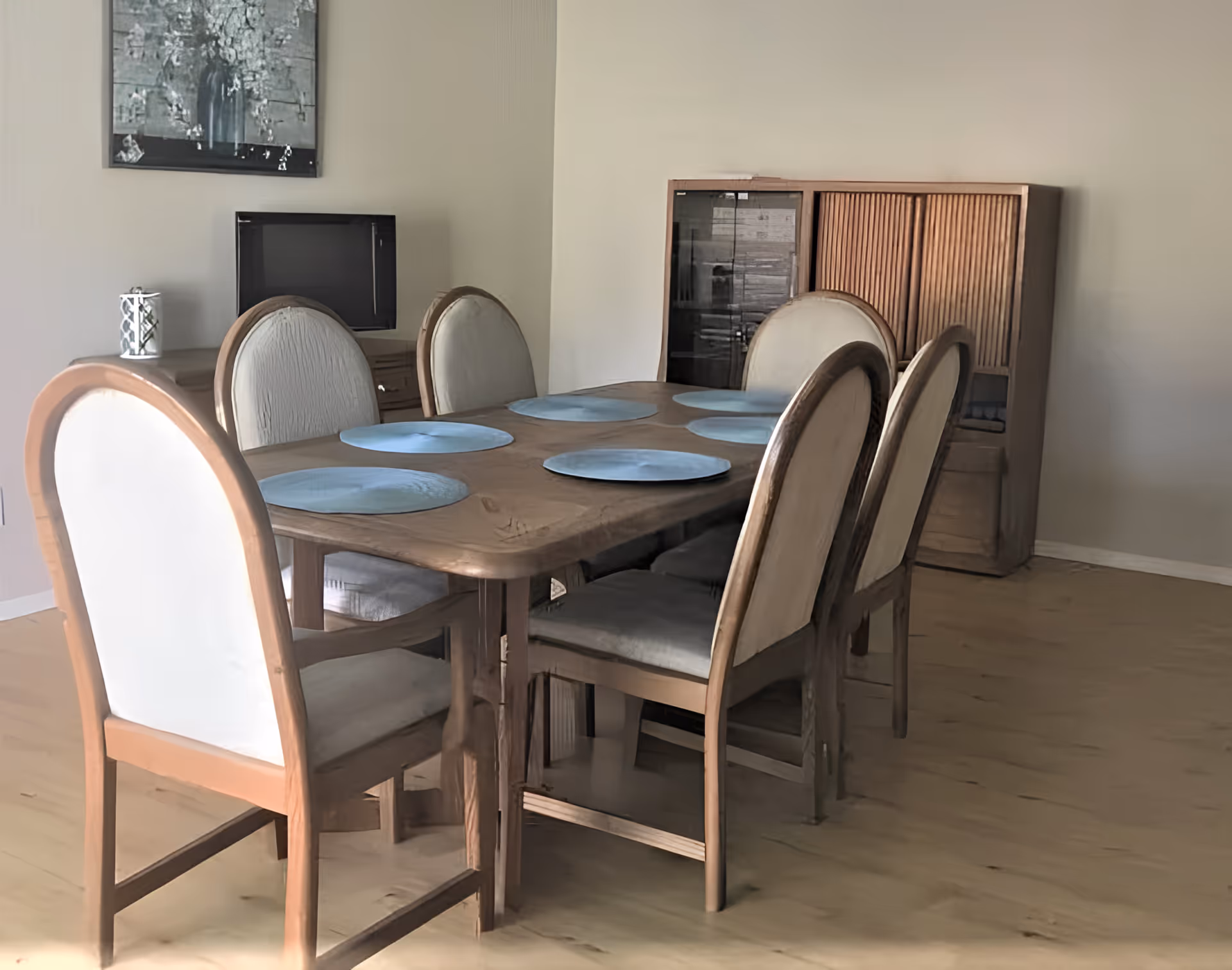 A dining room with a wooden table set with six light blue placemats and six cushioned chairs with wooden frames. In the background, there is a wooden cabinet with glass doors and a small TV on a stand. A framed picture hangs on the wall above the TV.