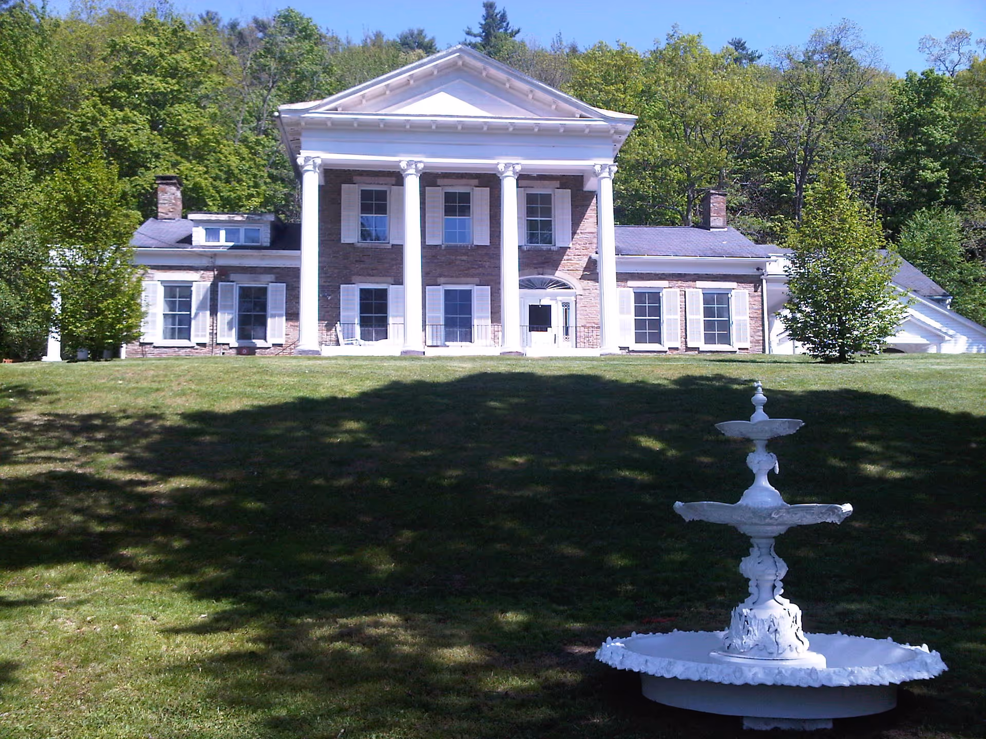 Front exterior view of a large, stately building with white columns and multiple windows, surrounded by green trees and a grassy lawn with a white decorative fountain in the foreground.