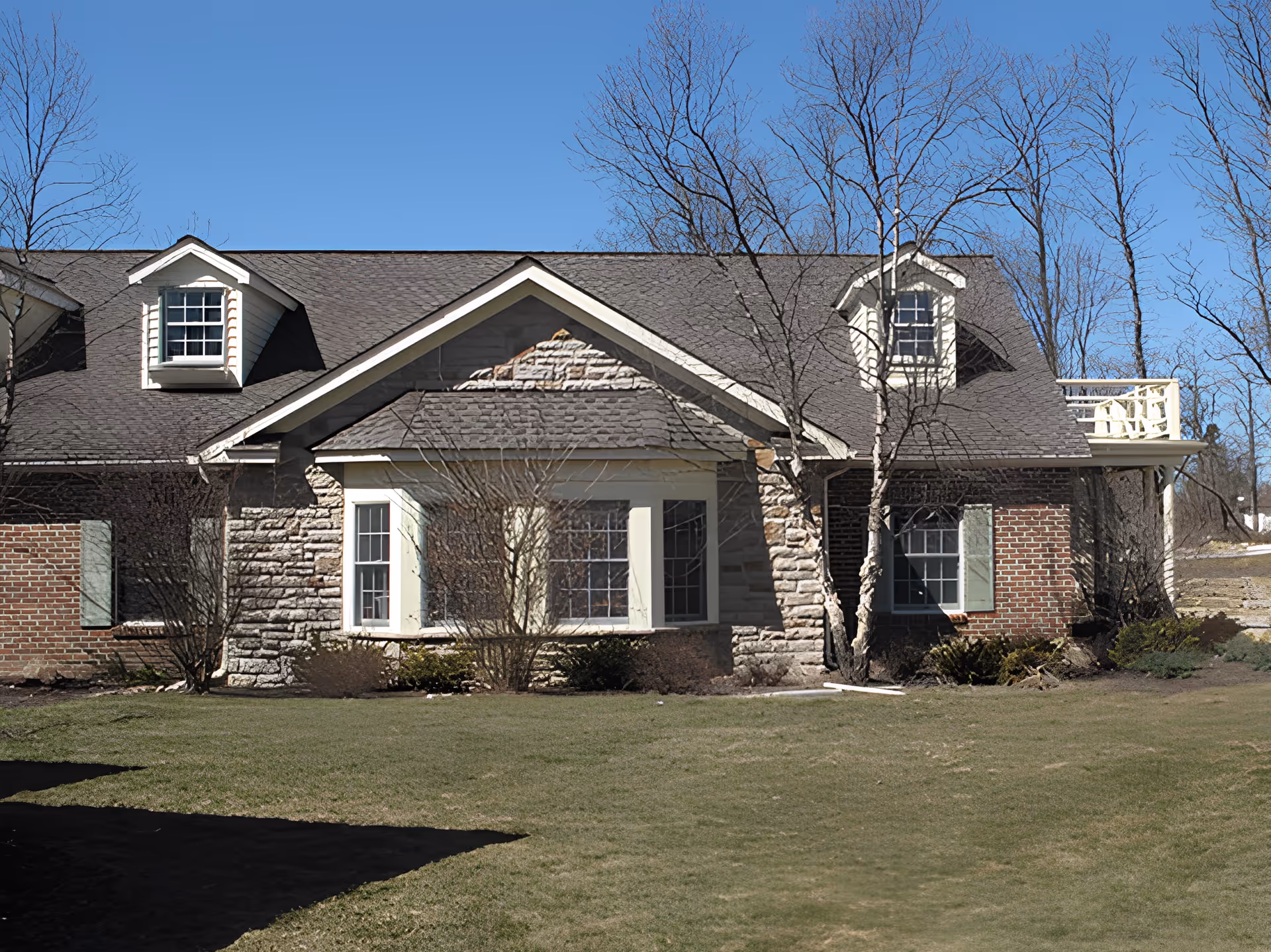 Exterior view of a single-story building with a combination of stone and brick walls, multiple windows with white frames and green shutters, and a dark shingled roof with dormer windows. Leafless trees and shrubs are in front of the building, and a grassy lawn extends in the foreground under a clear blue sky.
