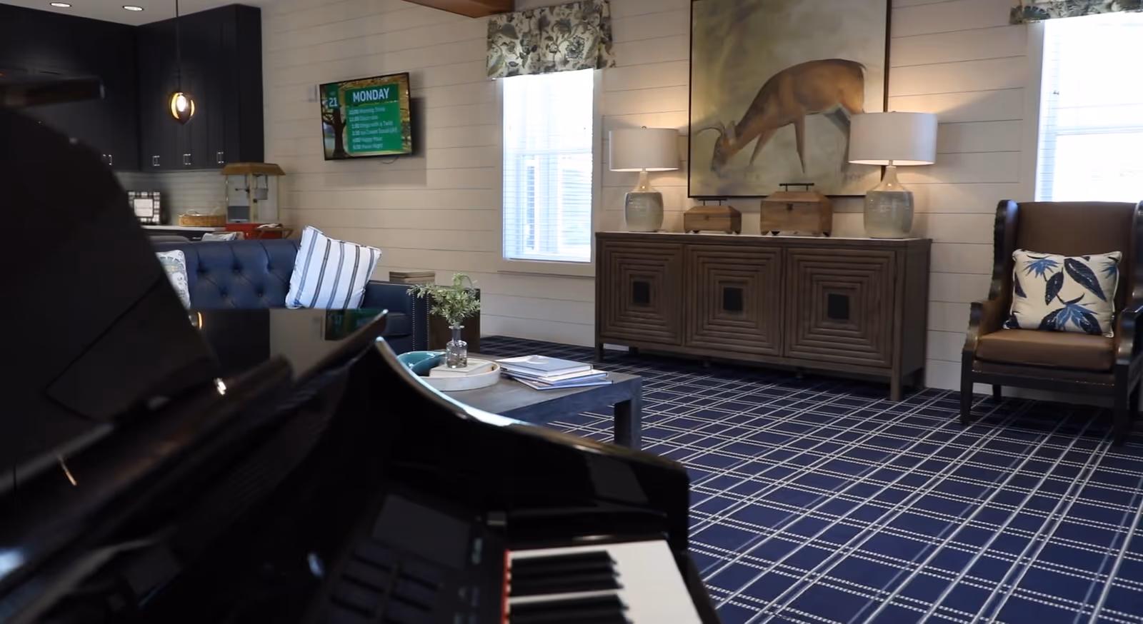 Interior view of a senior living facility common area featuring a black grand piano in the foreground, a blue tufted sofa with striped pillows, a wooden sideboard with two lamps and a large painting of a deer above it, a brown armchair with a floral cushion, and a blue and white checkered carpet. The room has white shiplap walls and two windows with floral valances.