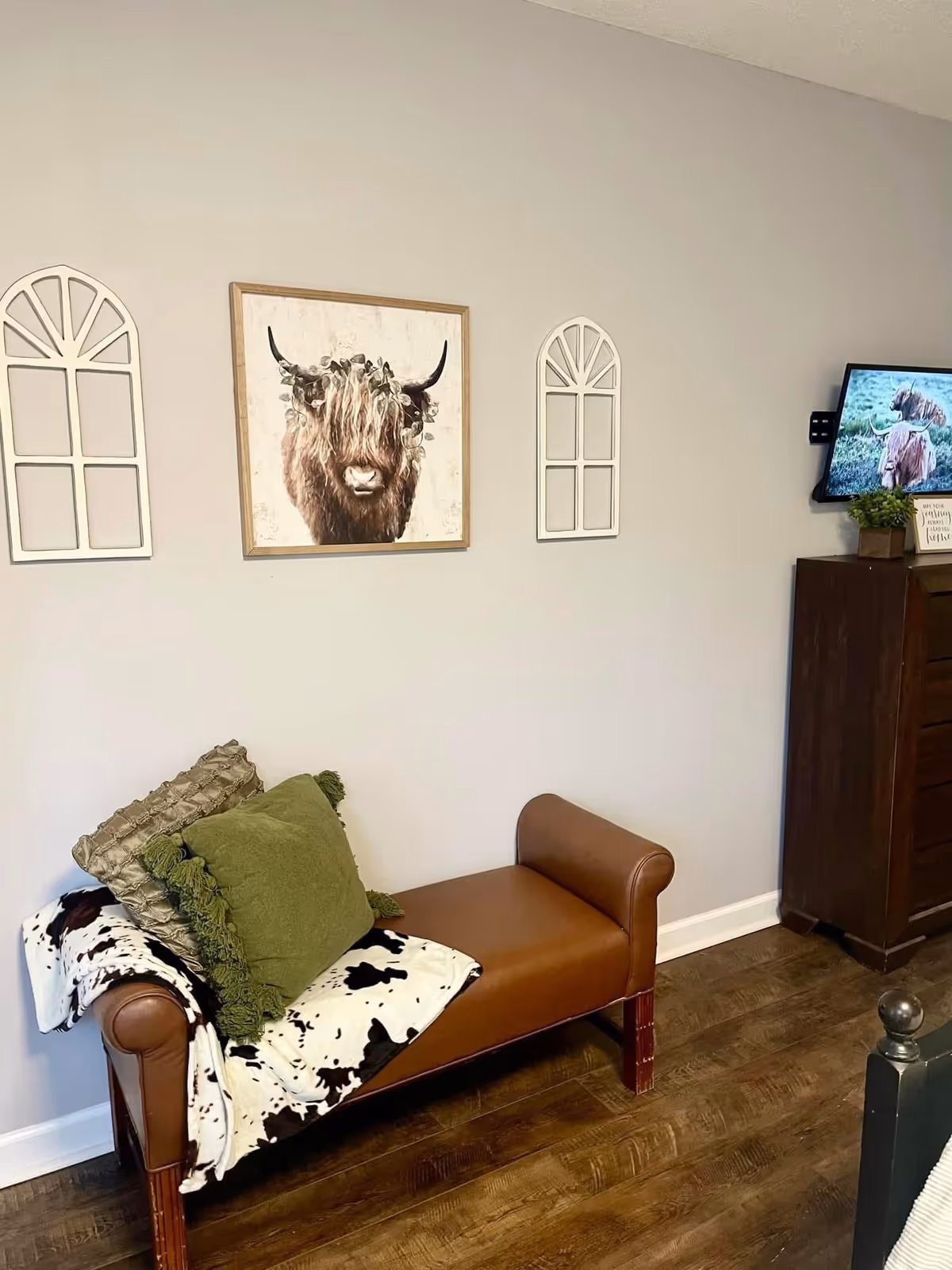 A small bedroom sitting area with a leather bench draped with a cowhide throw and pillows beneath framed wall art and a mounted TV.