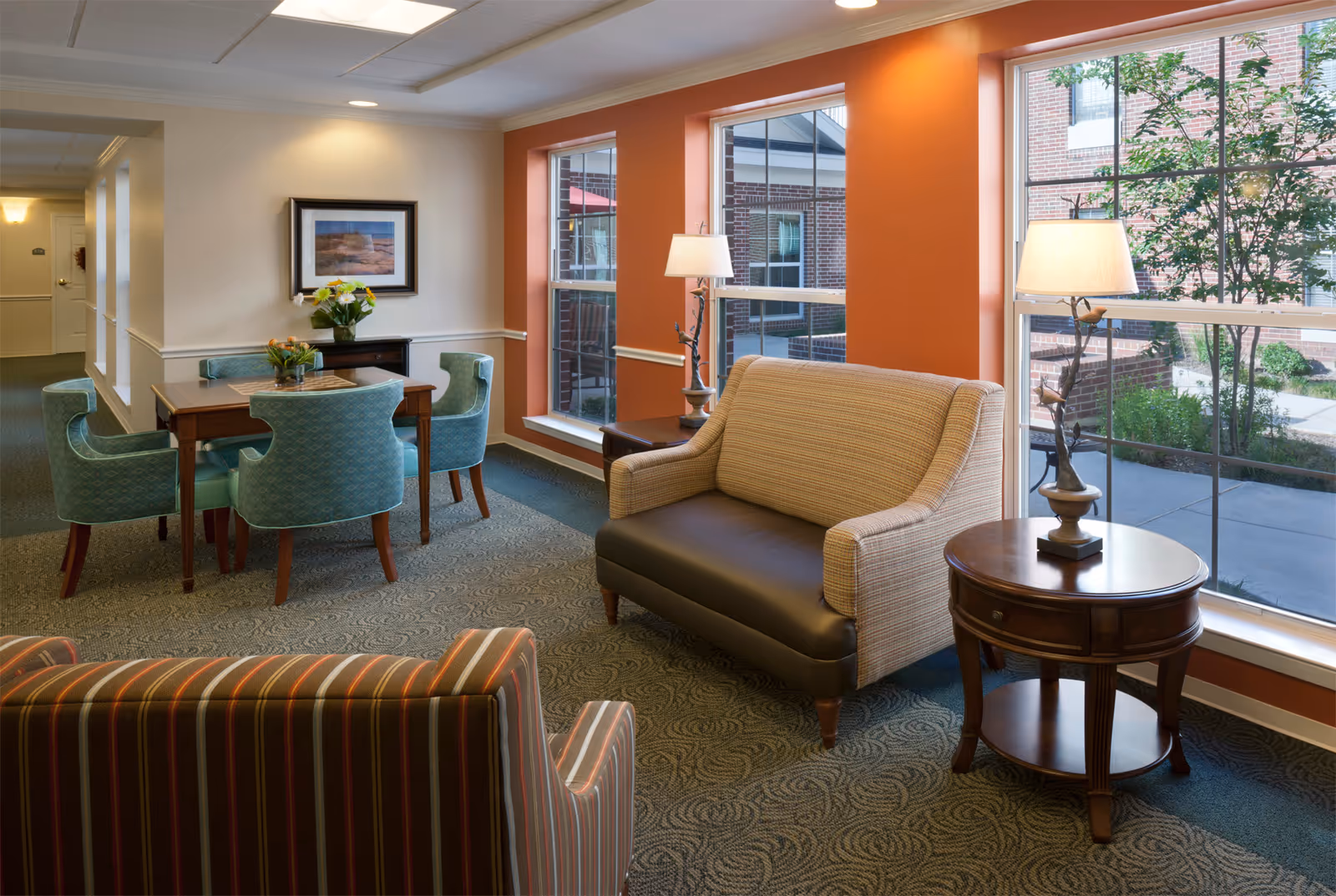 A cozy senior living common area with a small wooden table surrounded by four teal upholstered chairs, a striped armchair, and a beige loveseat with a dark brown seat cushion. The room features large windows with views of a brick courtyard and greenery outside. There are two wooden side tables with decorative lamps, and a framed picture on the cream-colored wall.