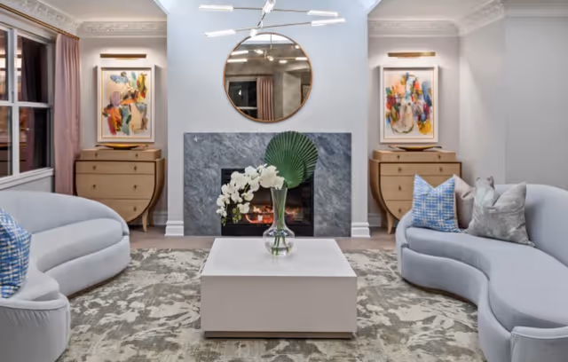 A modern living room with two curved light gray sofas facing each other, a white square coffee table with a glass vase holding white flowers and a large green leaf, a fireplace with a dark stone surround, a round mirror above the fireplace, two wooden dressers with abstract colorful paintings above them, and a patterned area rug on the floor.