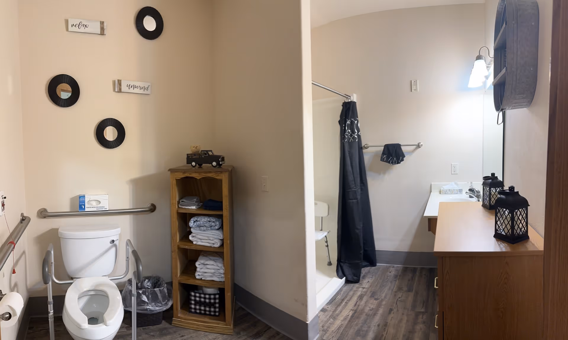 Accessible bathroom with a toilet and grab bars, a wooden shelf of towels, a curtained shower with a shower chair, and a sink vanity.