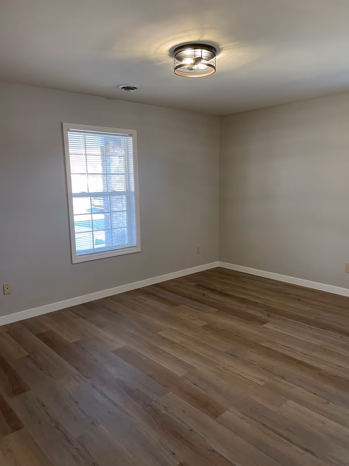Empty room with light wood flooring, a single window with blinds, beige walls, white baseboards, and a ceiling light fixture.