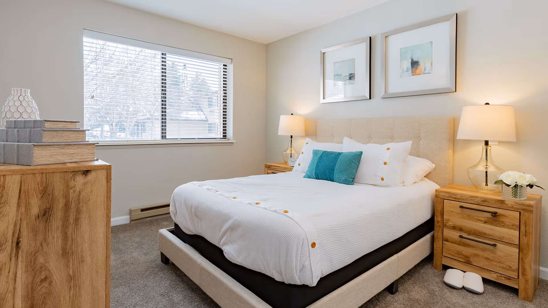 A cozy bedroom with a neatly made bed featuring white bedding and teal accent pillows. There are two wooden nightstands on either side of the bed, each with a lamp and one with a small vase of white flowers. A wooden dresser with stacked books and a decorative vase is positioned near a window with blinds, letting in natural light.