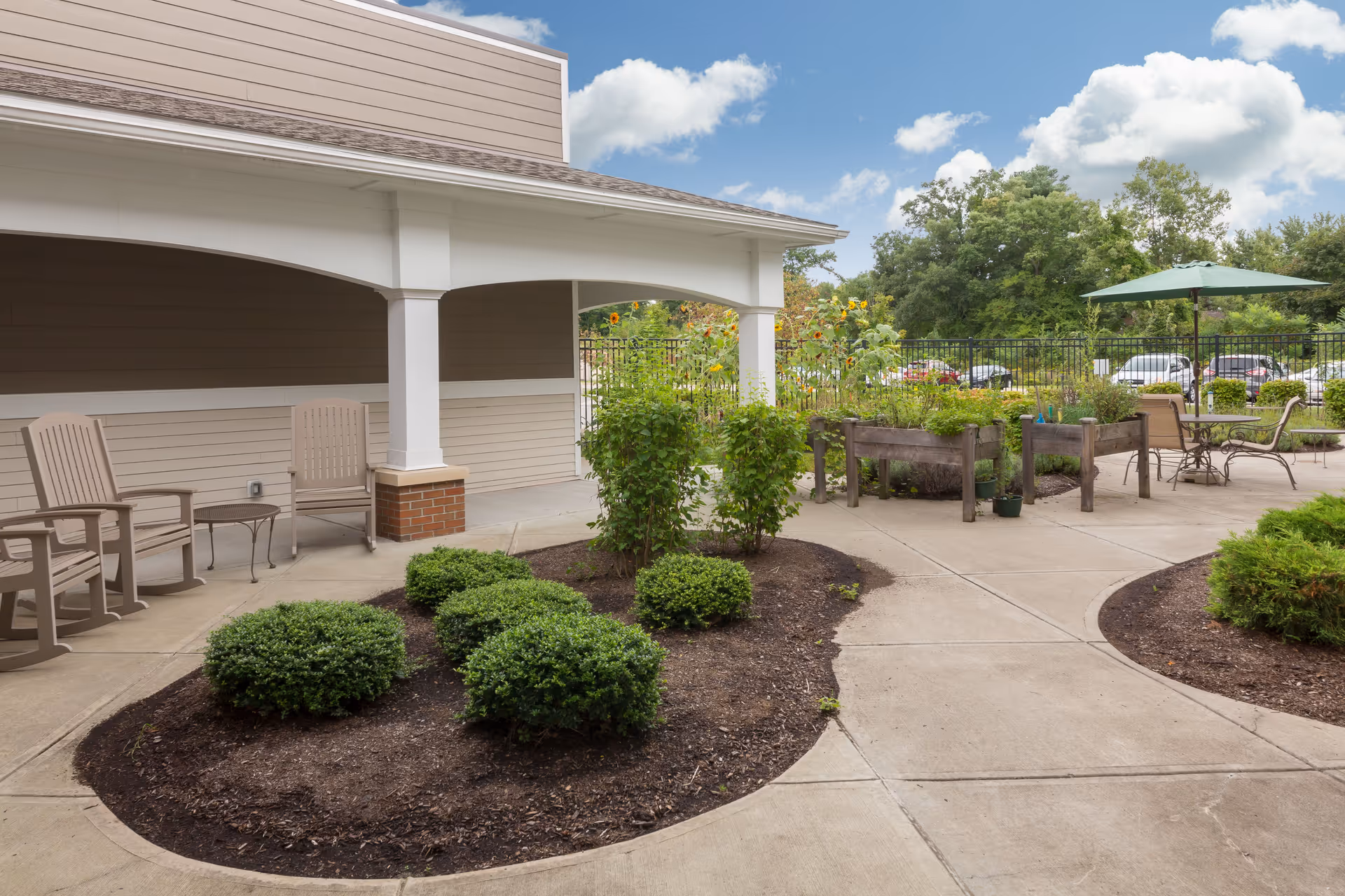 Outdoor patio area at The Reserve at East Longmeadow featuring a covered seating area with two chairs and a small table, landscaped garden beds with shrubs and plants, raised garden beds with greenery, and a patio table with chairs under a green umbrella. Trees and parked cars are visible in the background under a partly cloudy sky.