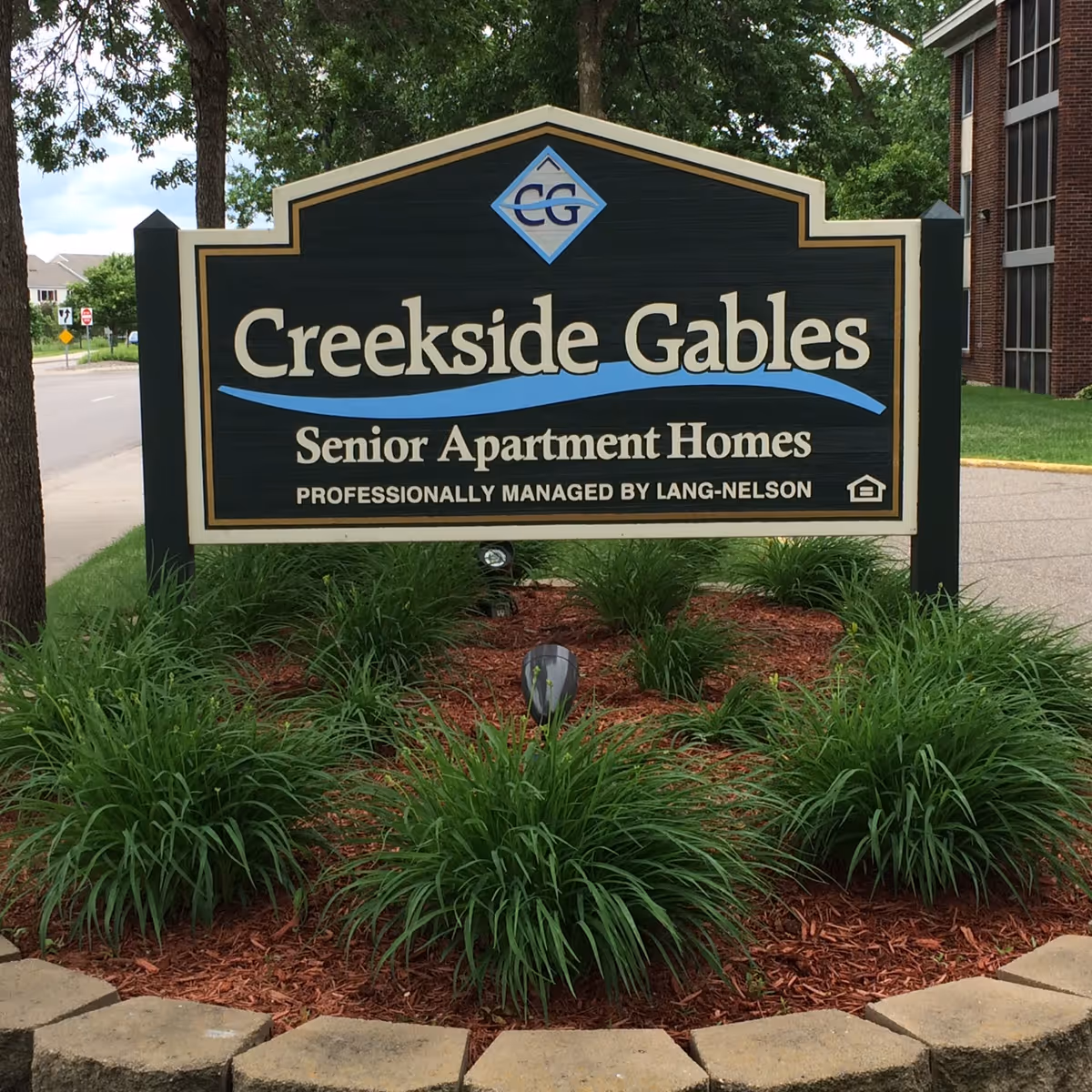 A large outdoor sign for Creekside Gables Senior Apartment Homes, professionally managed by Lang-Nelson, surrounded by green plants and mulch with a building and street visible in the background.