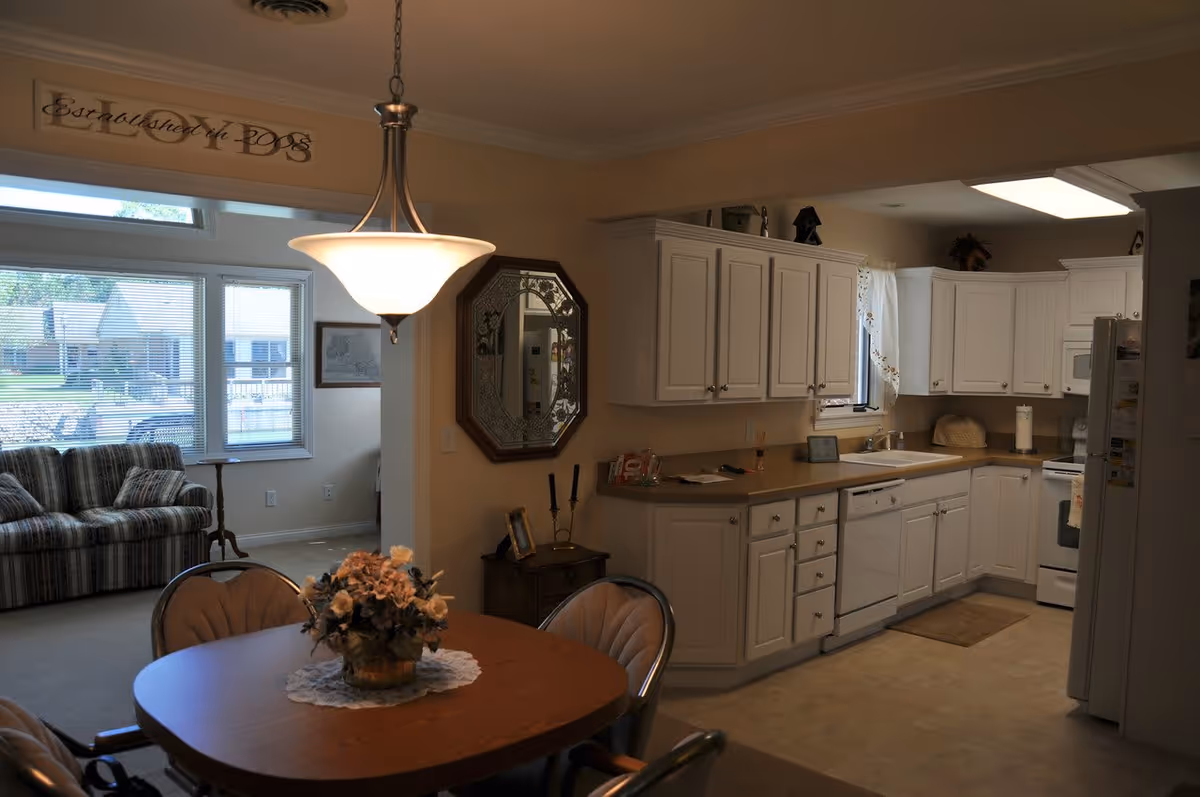 Interior view of a senior living community showing a kitchen and dining area. The kitchen has white cabinets, a dishwasher, stove, and refrigerator. The dining area has a wooden table with four chairs and a floral centerpiece under a hanging light fixture. In the background, there is a living room with a striped couch and large windows letting in natural light.