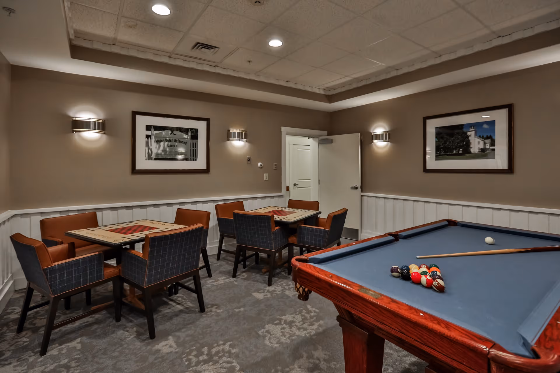 A recreational room with a pool table in the foreground and two tables with four chairs each in the background. The walls are beige with white wainscoting, and there are two framed pictures and wall-mounted lights. The ceiling has recessed lighting, and a door is open in the back wall.
