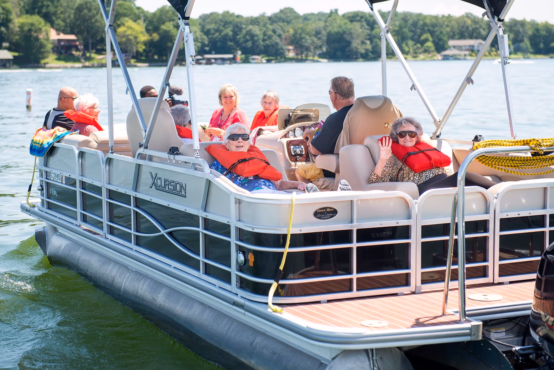 A group of elderly people wearing life jackets are sitting and relaxing on a pontoon boat on a lake, enjoying a sunny day with trees and houses visible in the background.