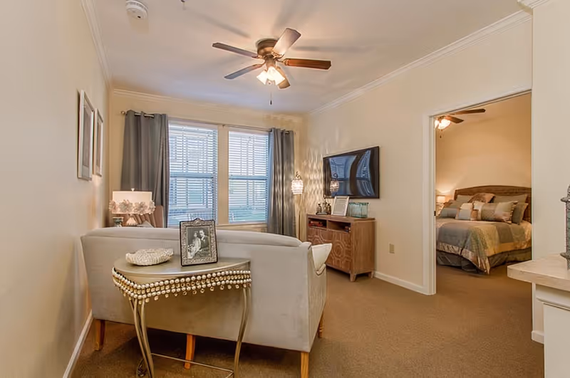 A cozy living room area with a beige couch facing a wall-mounted TV. There is a small decorative table behind the couch with a framed photo and a decorative bowl. The room has beige walls, carpeted floor, and a ceiling fan with lights. In the background, an open doorway leads to a bedroom with a bed made up with pillows and a quilt. The bedroom also has a ceiling fan and warm lighting.