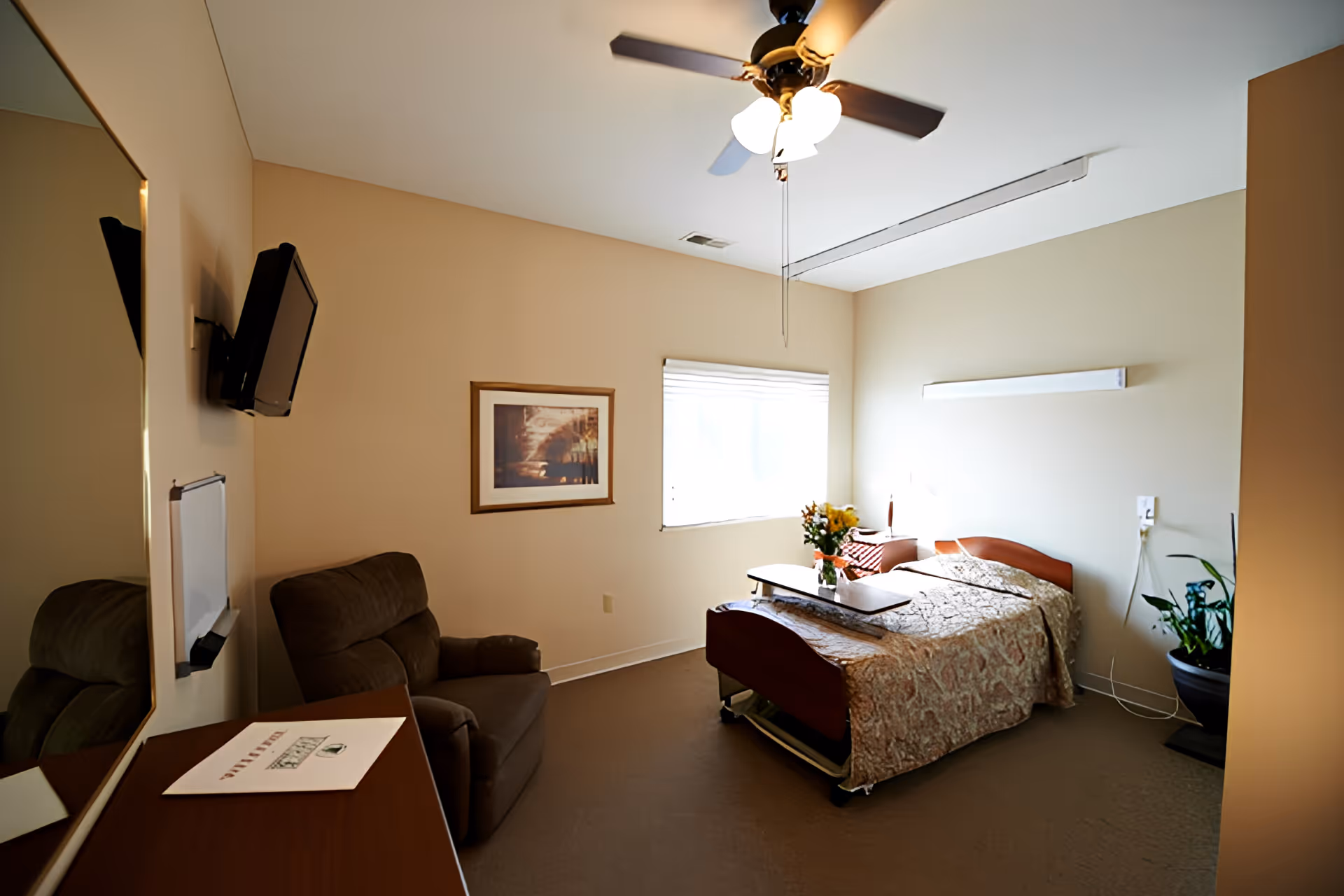 A single bedroom in a senior living facility with a bed covered in a patterned bedspread, a small table with flowers on it, a comfortable armchair, a wall-mounted TV, a framed picture on the wall, a window with blinds, a ceiling fan with lights, and a potted plant in the corner.
