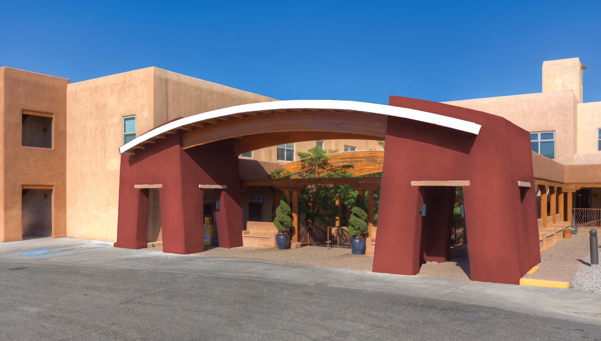 Exterior view of The Montecito of Santa Fe facility showing a southwestern-style building with beige stucco walls and a large red archway entrance with wooden beams and potted plants under a clear blue sky.