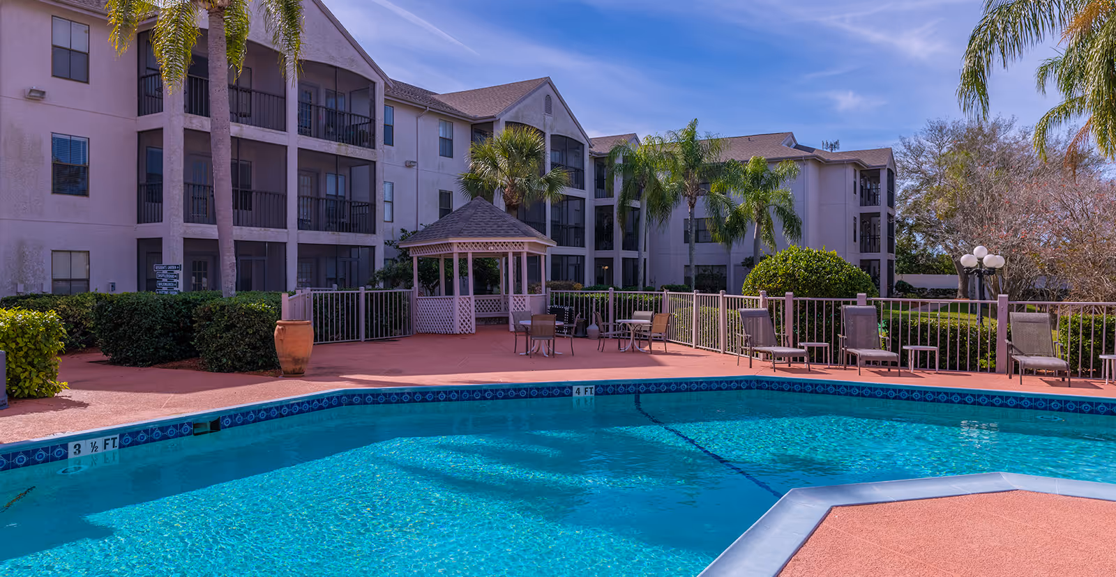 Outdoor swimming pool with lounge chairs, a gazebo, and a multi-story residential building with balconies under a blue sky.