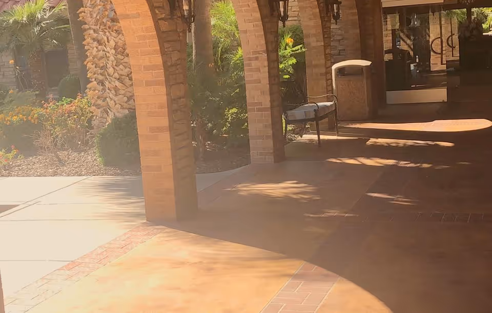 Outdoor covered walkway with brick arches and columns, a bench, and a trash can near the entrance door. There are palm trees and landscaped plants visible in the background.