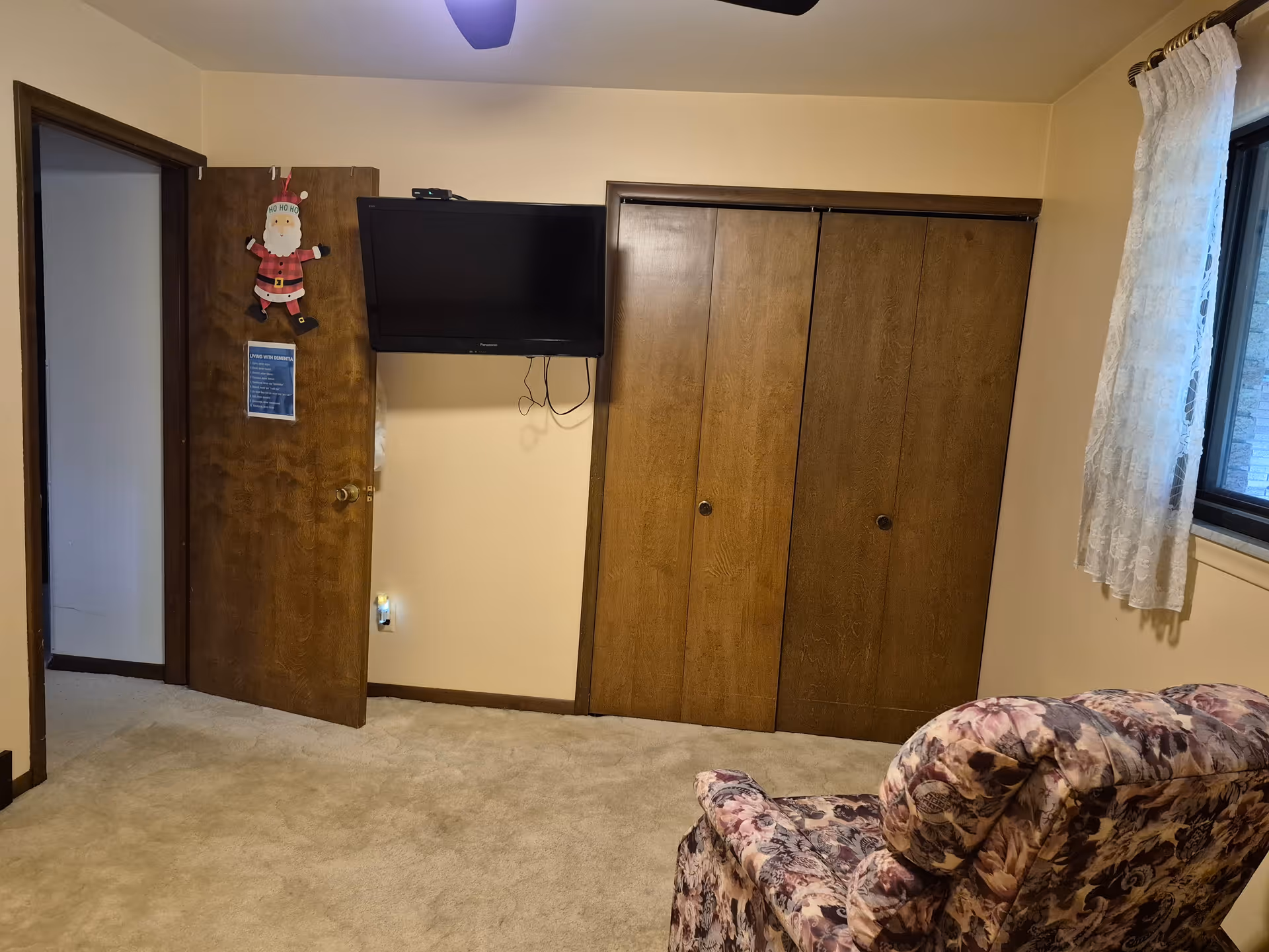 A room with beige walls and carpeted floor featuring a floral upholstered armchair in the foreground. On the far wall, there is a wooden door with a Santa Claus decoration and a sign, a wall-mounted flat screen TV, and a set of wooden closet doors. A window with white lace curtains is on the right side of the room.
