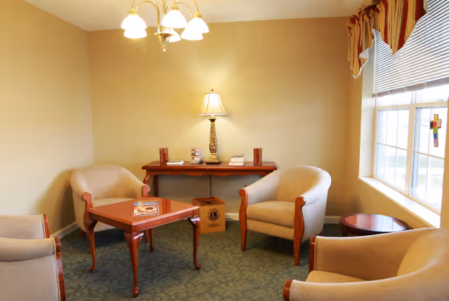 A cozy sitting area with four beige armchairs arranged around a wooden coffee table. Behind the chairs is a wooden console table with a lamp and several books. A window with blinds and a valance lets in natural light on the right side of the room. The walls are painted a light beige color and the floor is carpeted in a green patterned design.