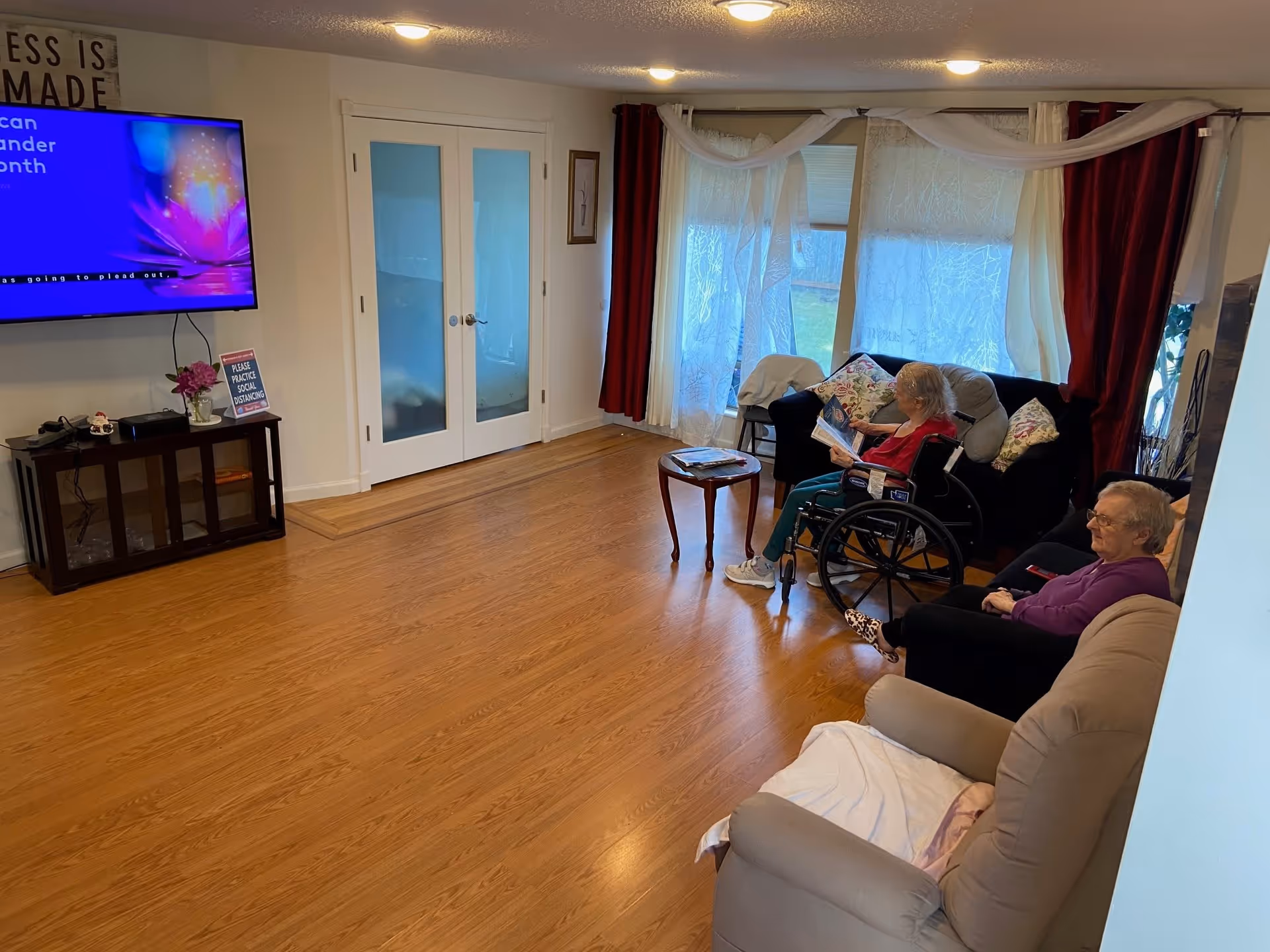 A living room with wooden flooring, a mounted flat-screen TV on the wall, a small cabinet with decorative items, and two elderly women seated on sofas. One woman is in a wheelchair reading a book, and the other is seated on a couch. The room has large windows with sheer white curtains and red drapes.