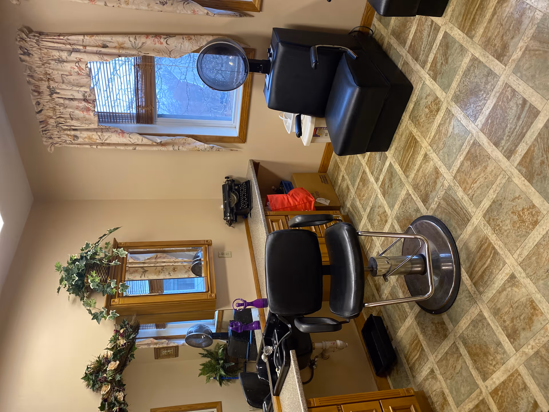 Interior of a salon room with a black salon chair in front of a counter with a sink, a mirror with a wooden frame, and decorative plants above. There is a window with floral curtains and a black hair dryer chair next to it. The floor has a patterned tile design.