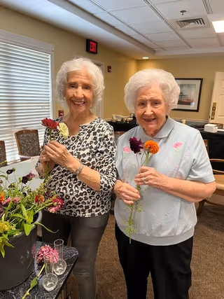 Two elderly women standing indoors holding colorful flowers and smiling. They are in a well-lit room with beige walls, a window with blinds, and framed artwork on the wall behind them. A table with more flowers and vases is visible in the foreground.