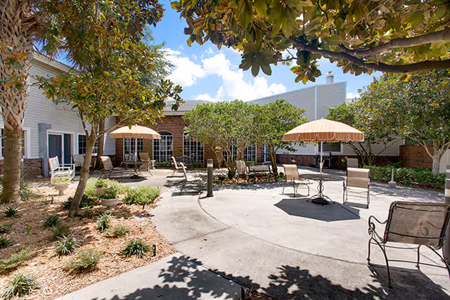 Outdoor patio area at Aldea Green with several tables and chairs under beige umbrellas, surrounded by trees and landscaping, adjacent to a brick and white building under a blue sky with some clouds.