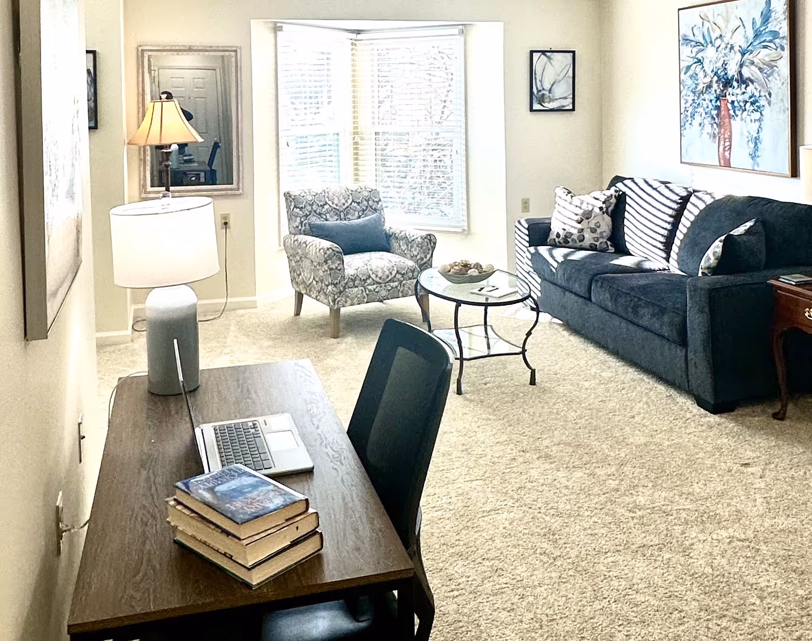 Bright living room with a desk and laptop in the foreground, a patterned armchair by a bay window, glass coffee table, and a dark sofa with pillows.