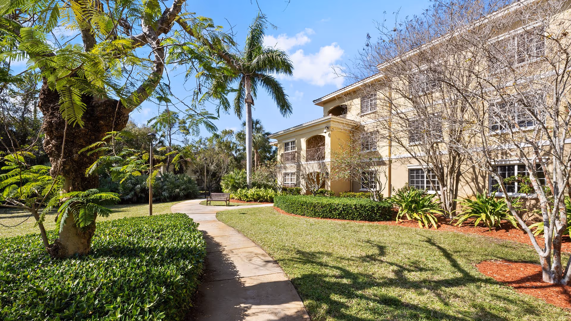 Outdoor view of a senior living facility with a paved walkway winding through a landscaped garden area featuring green bushes, palm trees, and other plants. A beige multi-story building with balconies and windows is visible on the right side under a clear blue sky.