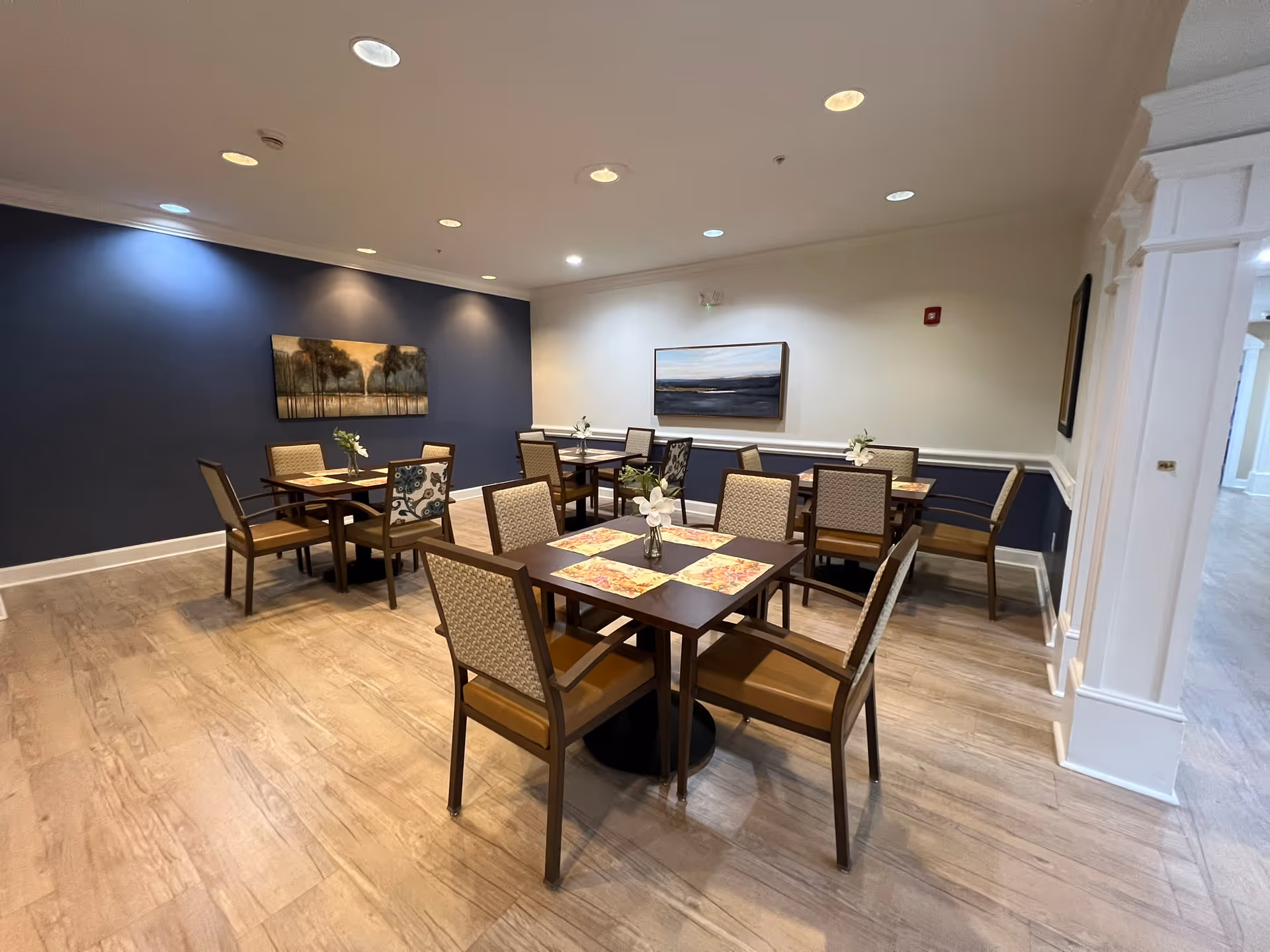 Well-lit dining room with several square tables and upholstered chairs, floral centerpieces, and artwork on navy and white walls.