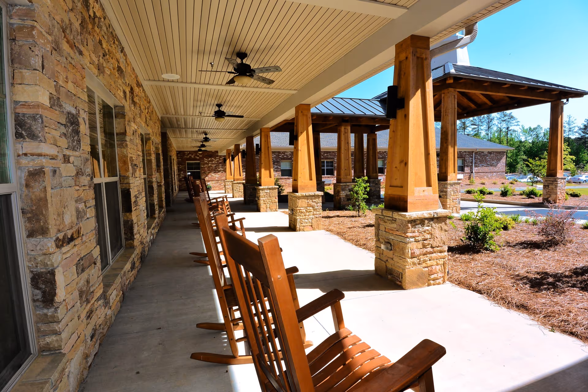 Covered outdoor porch area with wooden rocking chairs lined up along a stone wall with windows, wooden pillars supporting the roof, ceiling fans, and a landscaped garden area with shrubs and mulch under a clear blue sky.