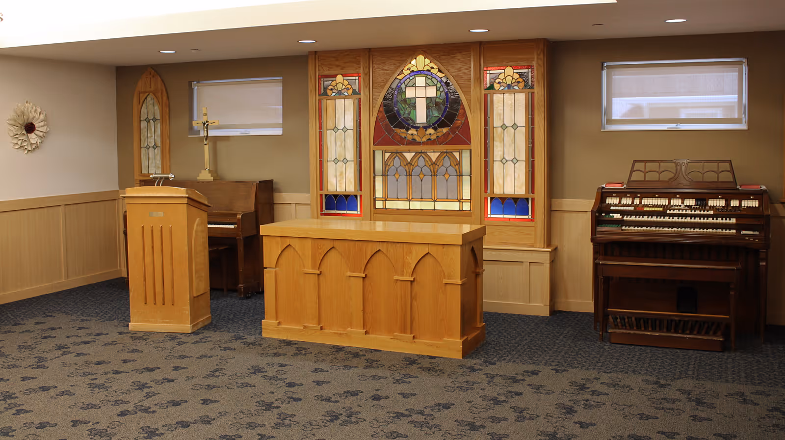 Interior room with a wooden altar, a wooden podium, a piano, and an organ. The back wall features a stained glass window with a cross design. The room has carpeted flooring and beige walls with wood paneling.