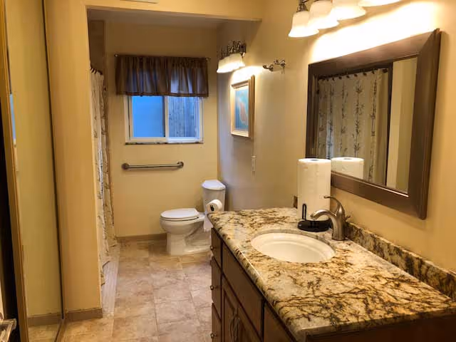 A clean bathroom with a granite countertop vanity featuring a sink, faucet, and two rolls of paper towels. There is a large mirror above the vanity and two light fixtures with three bulbs each. The bathroom has a toilet with a grab bar on the wall behind it, a window with a brown valance, and a shower area with a curtain on the left side.