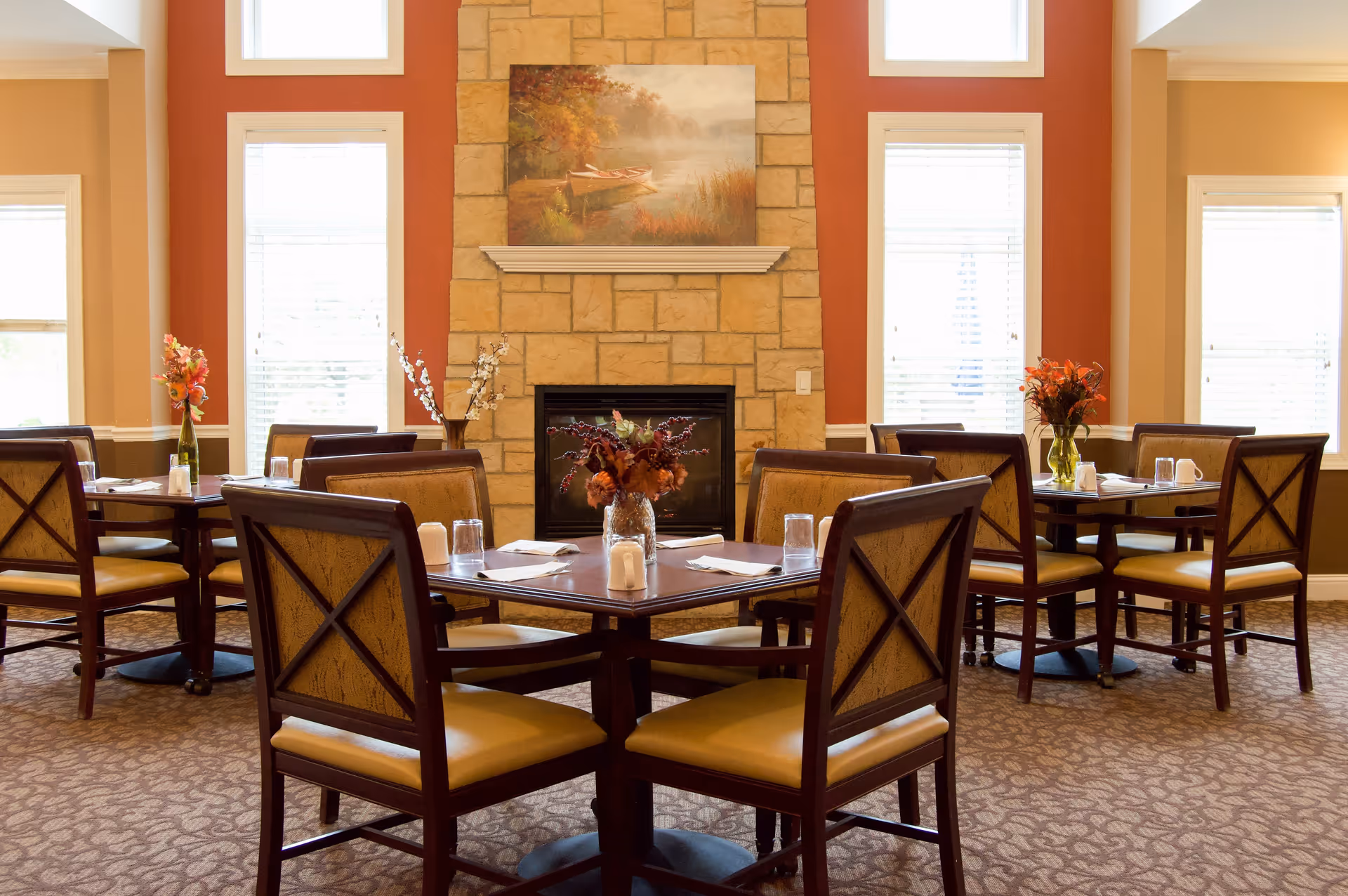 A cozy dining room with several wooden tables and cushioned chairs arranged around them. Each table has a small floral centerpiece and place settings with glasses and napkins. The room features large windows with white blinds, a stone fireplace with a painting of a boat on a lake above it, and warm-toned walls.