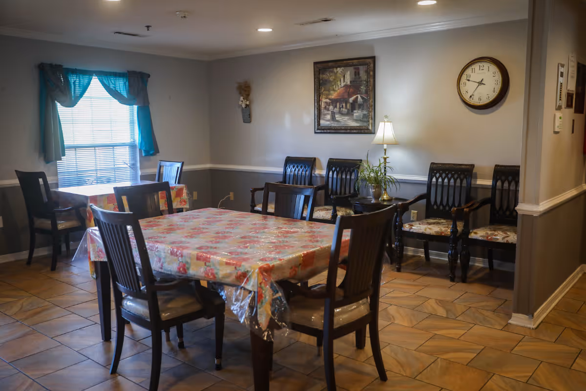 A dining room with two tables covered in floral-patterned plastic tablecloths, surrounded by wooden chairs. There is a window with blue curtains on the left wall, a framed painting and a wall clock on the right wall, and a small table with a lamp and a plant between several wooden chairs with patterned cushions.