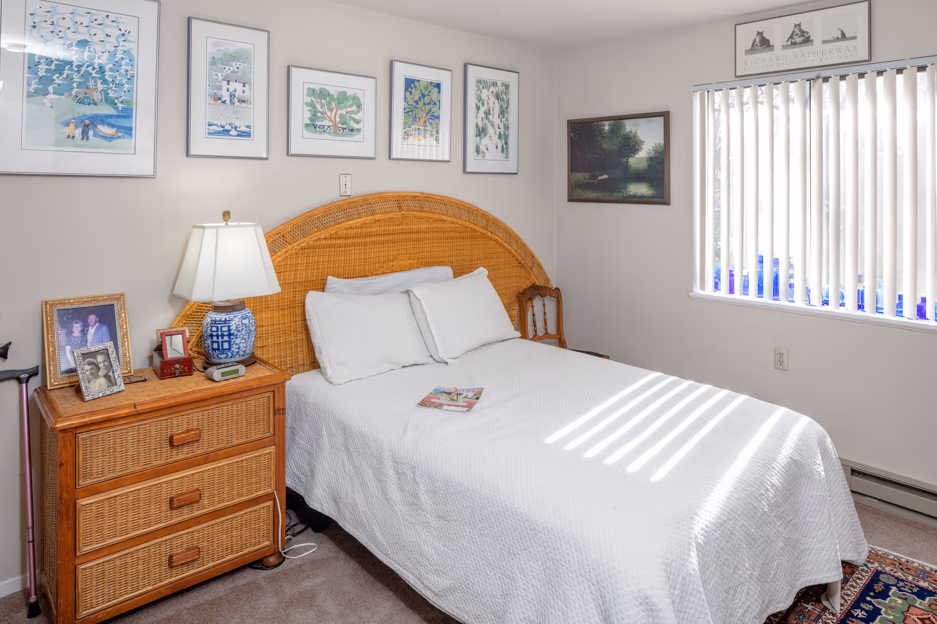 A cozy bedroom with a wicker headboard bed covered in a white quilt and two pillows. A wooden nightstand with three drawers sits beside the bed, holding a blue and white lamp, framed photographs, and a small clock. Above the bed, five framed artworks are hung on the wall. A window with vertical blinds lets in natural light, casting shadows on the bed. A small wooden chair is placed near the window.